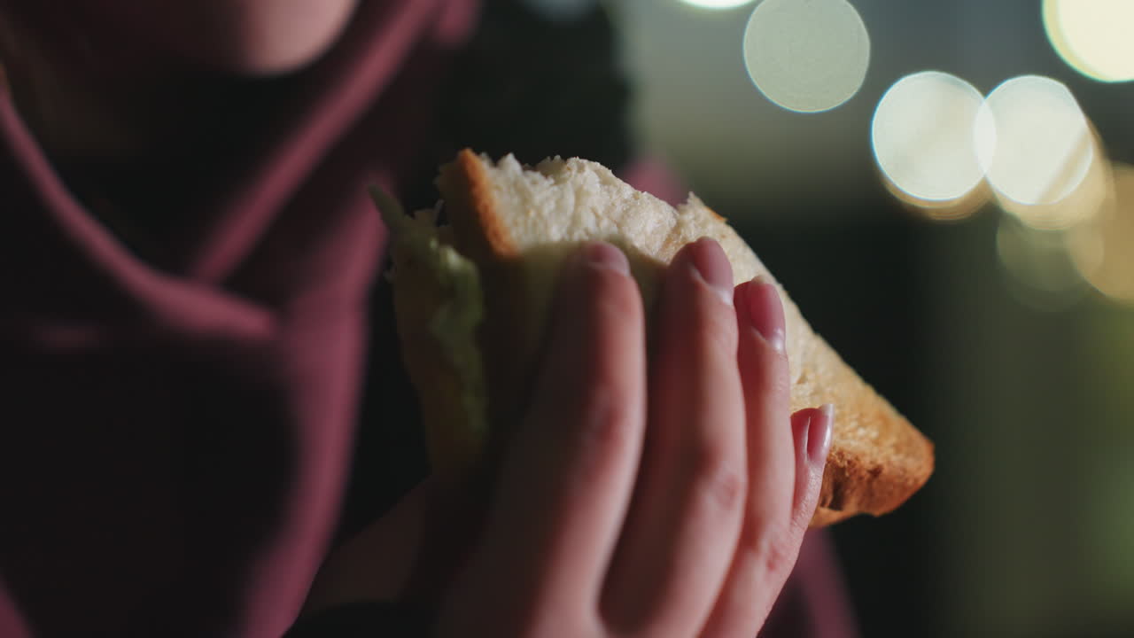Beautiful lady gently munching on bread sandwich outdoors on bench at twilight under blurred city lights capturing calm reflective moment and nourishing post workout break in urban night scene