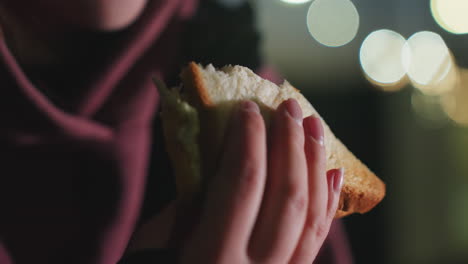 beautiful lady gently munching on bread sandwich outdoors on bench at twilight under blurred city lights capturing calm reflective moment and nourishing post workout break in urban night scene