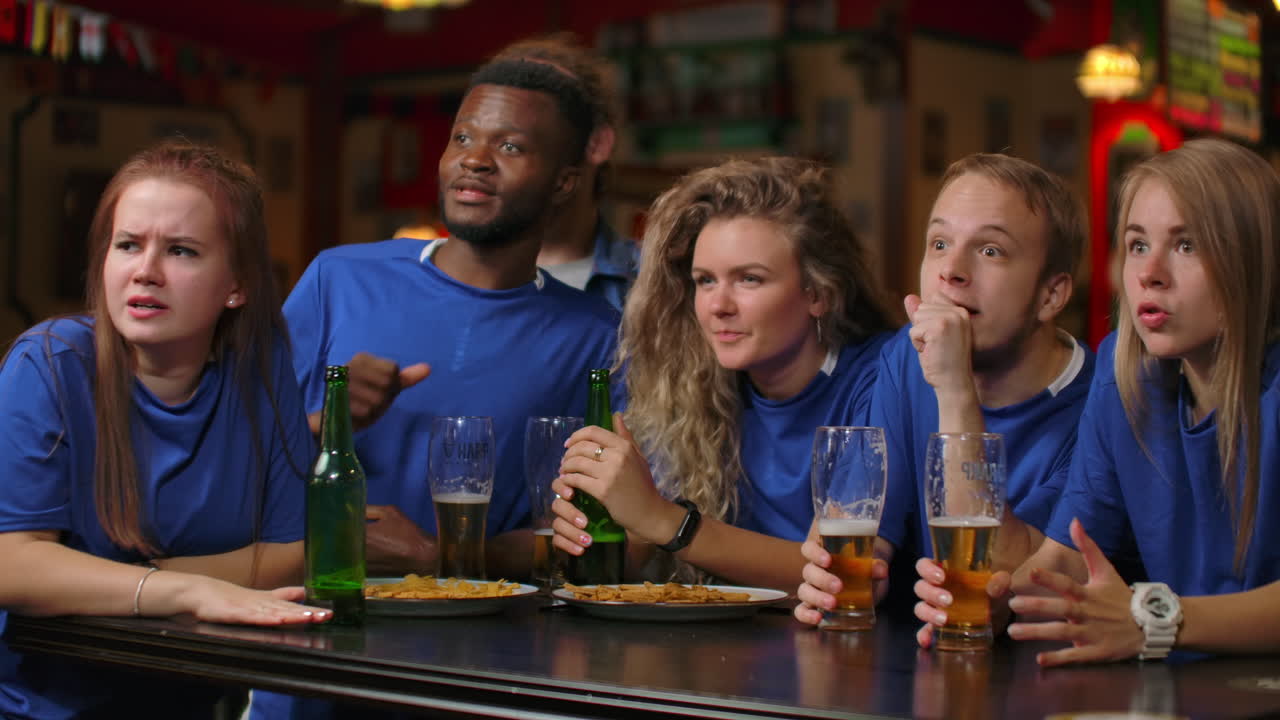Un gruppo di tifosi, uomini e donne in magliette blu, esultano urlando e bevendo birra al bar. Afroamericani ed europei