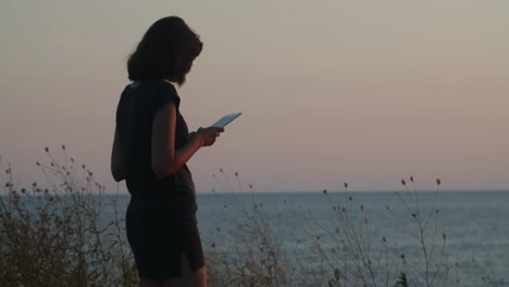 Chica-Parada-En-Un-Acantilado-Junto-Al-Mar-Al-Atardecer,-Con-Una-Tableta-En-Las-Manos,-Una-Toma-Más-Amplia