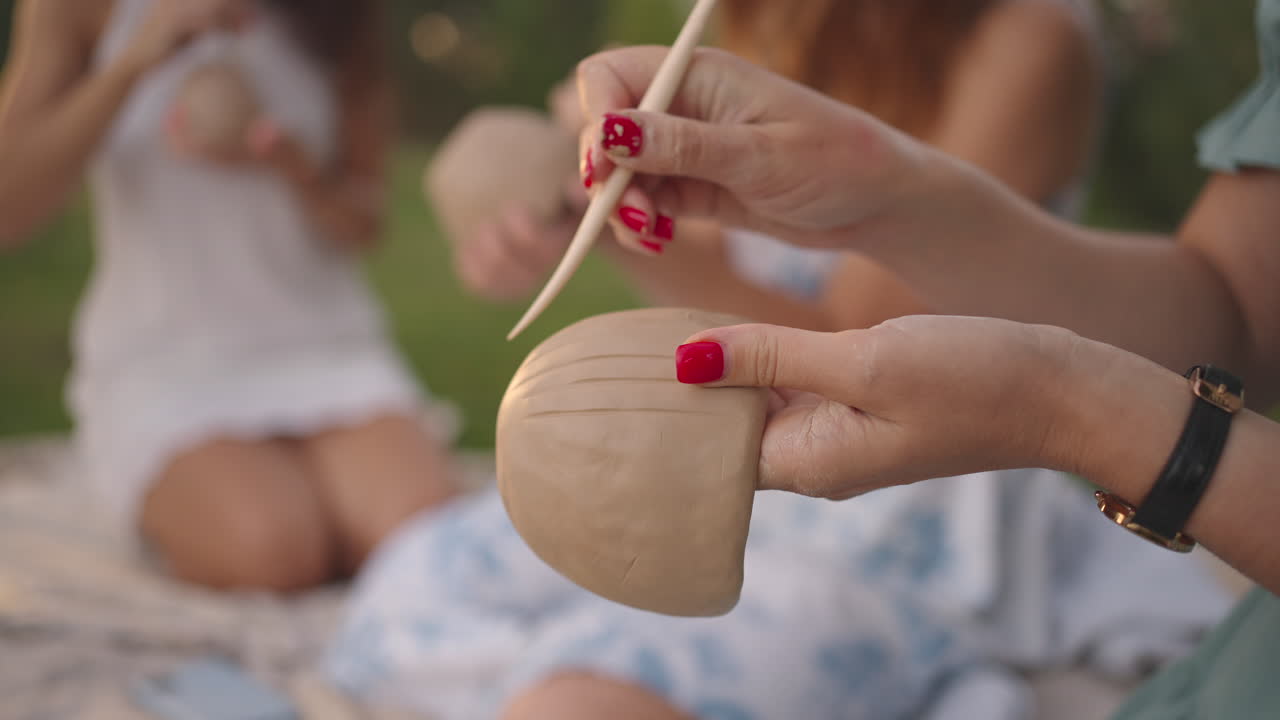 A group of young women are concentrating on applying patterns to clay products with the help of tools in a meadow in nature in an open space. Women's hands decorate the product in close-up.