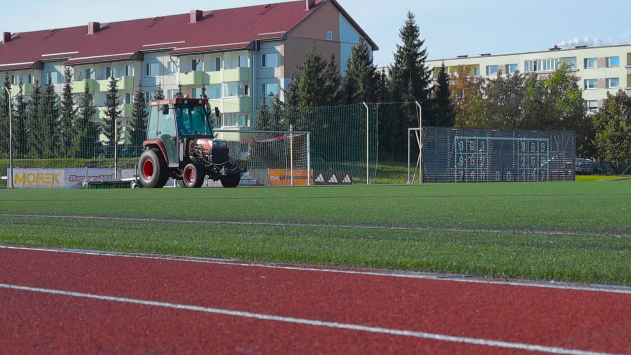 Small red tractor working on a football or a soccer stadium field maintaining it with dragging some equipment behind it to clean the green stadium ground during sunny autumn or summer day, urban shot.