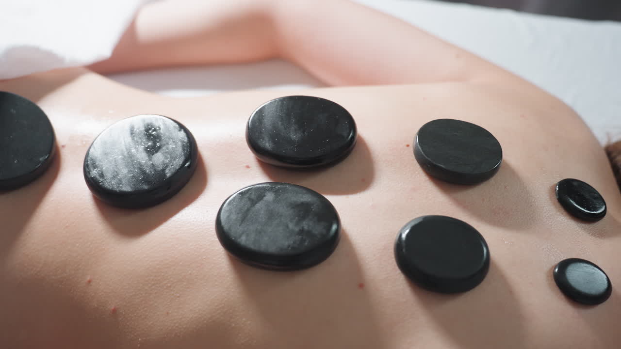 Close view of client with blond hair lying face down during calming spa treatment as seven smooth black massage stones are carefully arranged along bare back under soft natural light