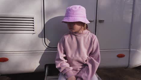 Portrait-of-a-blonde-girl-in-a-pink-hat-sitting-on-the-outside-steps-of-a-caravan