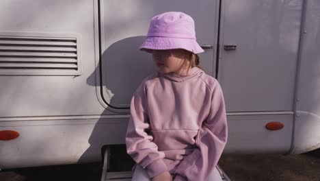 portrait of a blonde girl in a pink hat sitting on the outside steps of a caravan