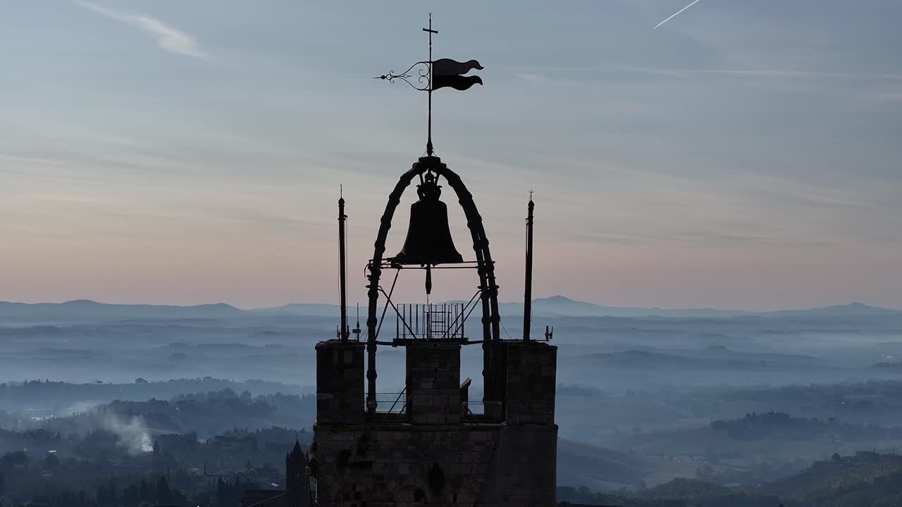Silhouette of a medieval bell tower and weathervane against the morning sky over foggy Tuscan rolling hills and valleys