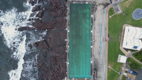 Große-Wellen-Krachen-In-Der-Nähe-Eines-Gemeinschaftsschwimmbads-An-Der-Küste,-Während-Menschen-Sport-Treiben