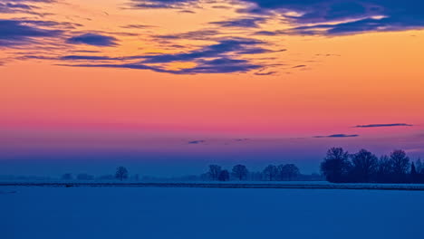 Vista-De-5k-De-Hermosos-Tonos-Naranja-Y-Amarillo-Después-De-La-Puesta-De-Sol-Y-Llanuras-Cubiertas-De-Nieve-Con-Pocos-árboles-En-Una-Noche-Invernal