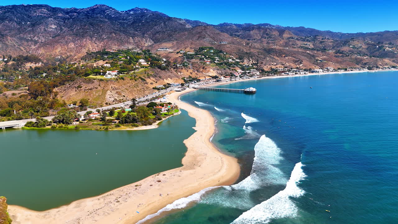 Empty line of a sandy beach with waves slowly rolling to. Houses scattered on the mountains. Malibu, Los Angeles County, California, USA. Aerial view