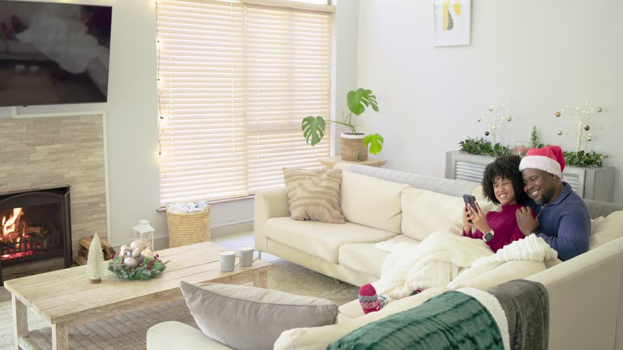 Diverse couple snuggling on sofa with festive decor, scrolling phone browsing content, copy space