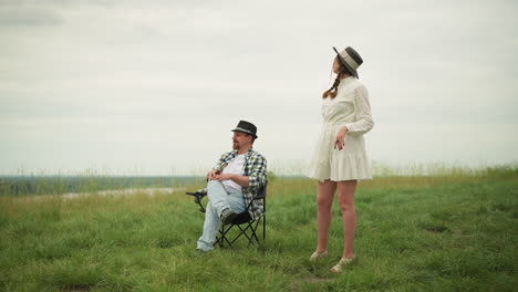 an artist in a checked shirt sits on a chair in a grassy field, holding brushes and crossing his leg to demonstrate a sitting pose to a woman in a white dress. the woman watches as he guides her