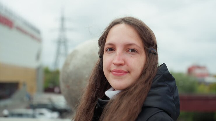 Donna bianca sorridente su un ponte cittadino, studentessa con capelli lunghi che si guarda intorno, appoggiata a una balaustra di pietra con monumento sferico dietro, cielo nuvoloso, traliccio dell'alta tensione ed edifici suburbani sullo sfondo
