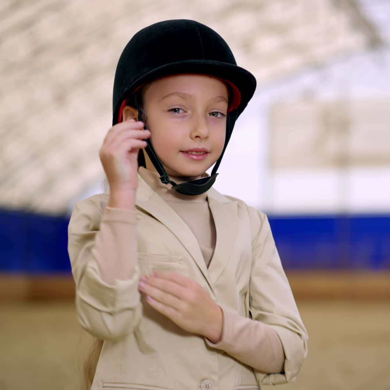 Young confident horse rider in black jockey helmet stands holding hands folded on her chest. Girl touché her hat looking at camera. Close up. Blurred backdrop