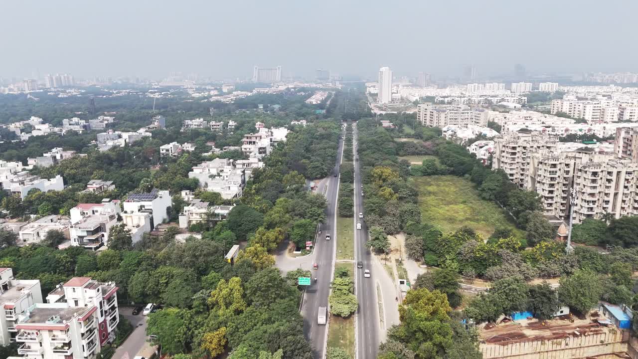 Un lussureggiante corridoio verde attraversa lo skyline residenziale di Greater Noida, dove viali alberati e complessi residenziali di media altezza si estendono in una foschia attenuata dall'inquinamento atmosferico urbano.