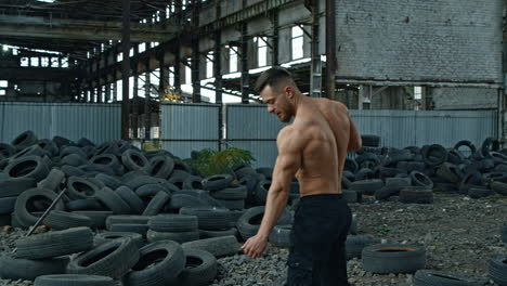 shirtless bodybuilder in abandoned place. young athlete with trained muscles posing to camera on used car tires background.