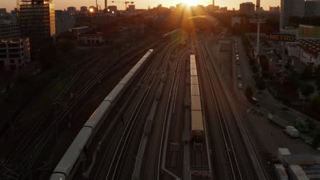 Beautiful-Establishing-Shot-of-white-passenger-Train-on-driving-into-Sunset-over-Berlin,-Germany-Golden-Hour-Cityscape-and-Ostbahnhof-Central-Train-Station,-Aerial-Wide-Angle