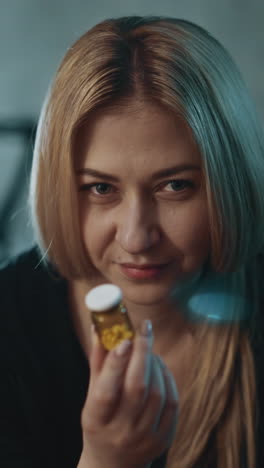 blonde woman with inscrutable smile holds small pill bottle of new nutritional supplement near workplace in semi-dark office room close view slow motion