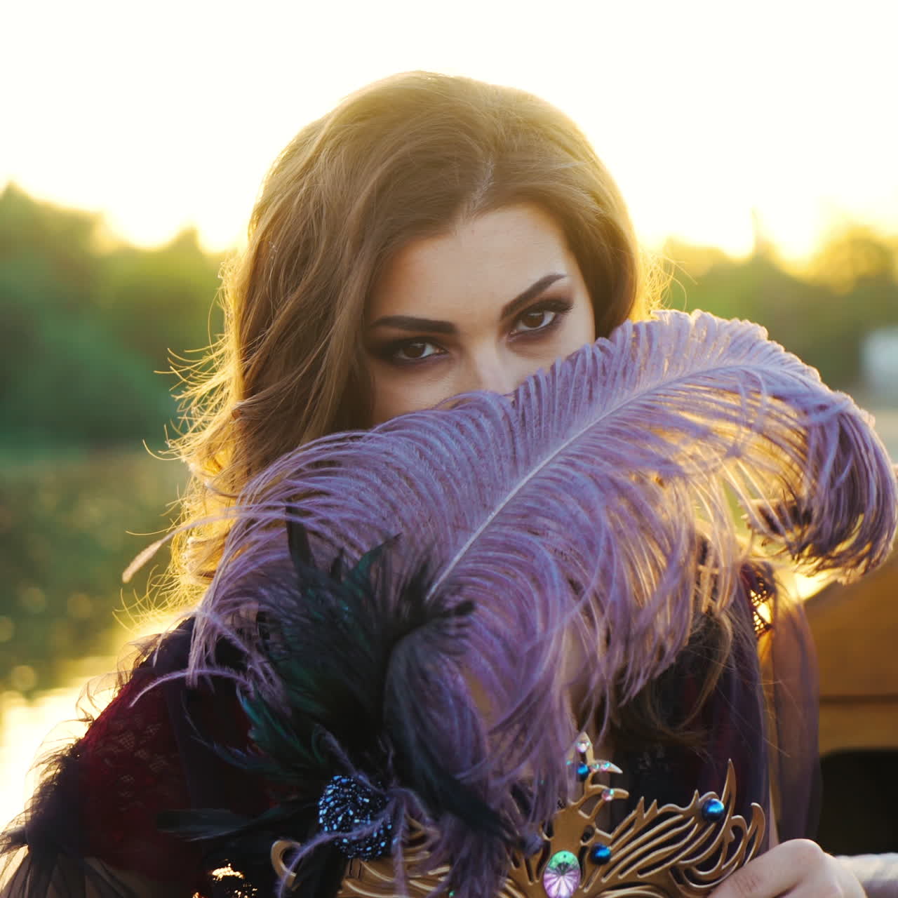 Beautiful woman posing into a camera while floating in a boat and closing her face with feather at sunset. Pretty female holding carnival mask with purple feather. Close-up