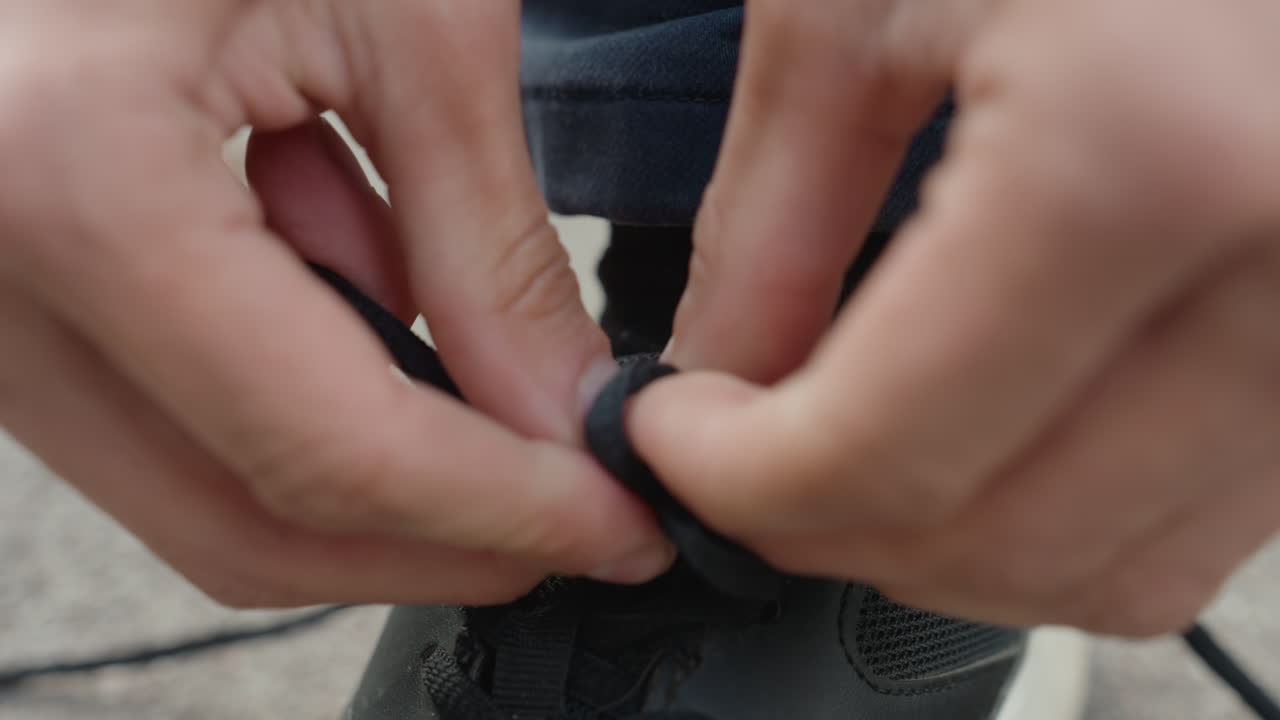 Close up tangan pria mengikat tali sepatu menjadi simpul yang aman, jari-jari fokus menarik ujung tali dan mengencangkannya, ujung celana jeans dan kaus kaki terlihat, teknik ini menunjukkan ketangkasan dan rutinitas pelajar, atlet, komuter
