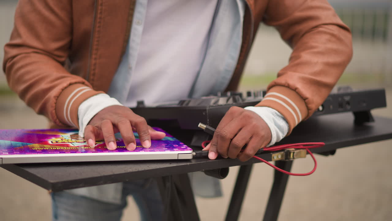 hands mixing on dj controller outdoors, red cable plugged into mixer, colorful record sleeve on table, leather jacket and white shirt visible, focused gestures over faders and knobs, urban street