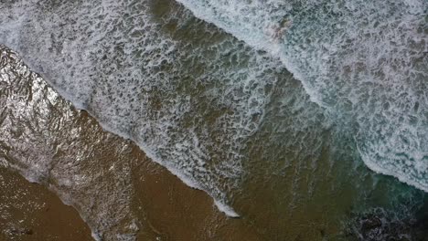 Toma-Aérea-Ascendente-Con-Un-Dron-Sobre-La-Costa-Que-Muestra-Las-Tranquilas-Olas-Turquesas-Y-La-Espuma-Blanca-Sobre-La-Arena-Naranja-Y-El-Brillo-Del-Reflejo-Del-Sol-En-Cantabria,-España.