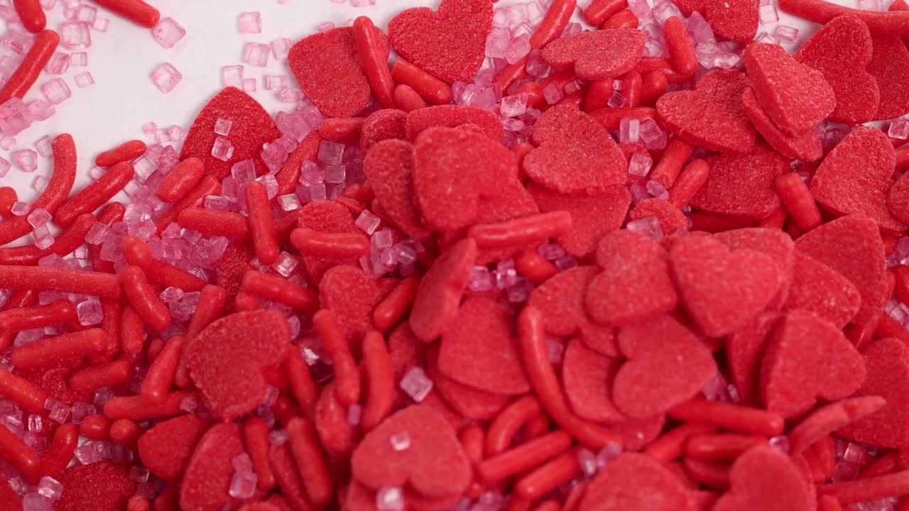Red heart-shaped candies and sprinkles cascade onto a surface in a vibrant, close-up slow-motion sequence