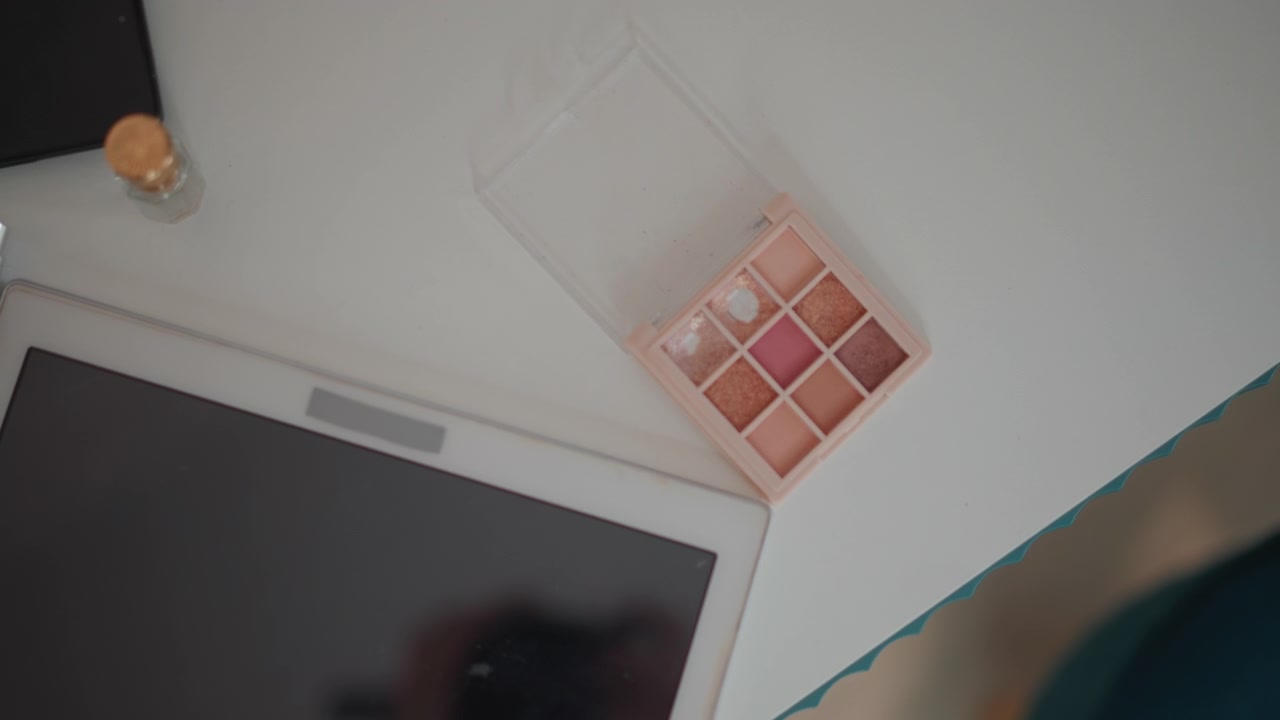 Overhead view shows arm reaching across bright white desk toward tilted tablet beside open eyeshadow palette with visible color pans, under soft daylight and gentle shadows in clean minimal workspace