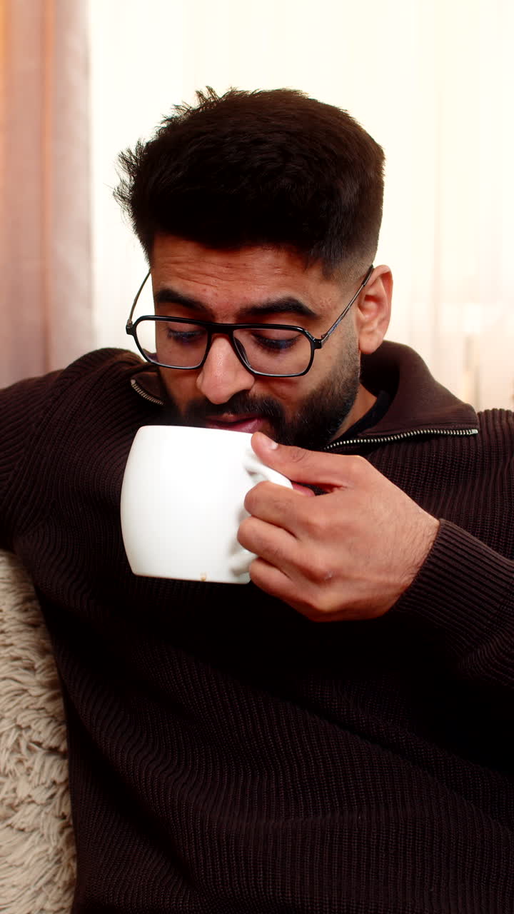 Indian man enters room on sofa enjoys coffee with a smile after stressful day at home with comfort