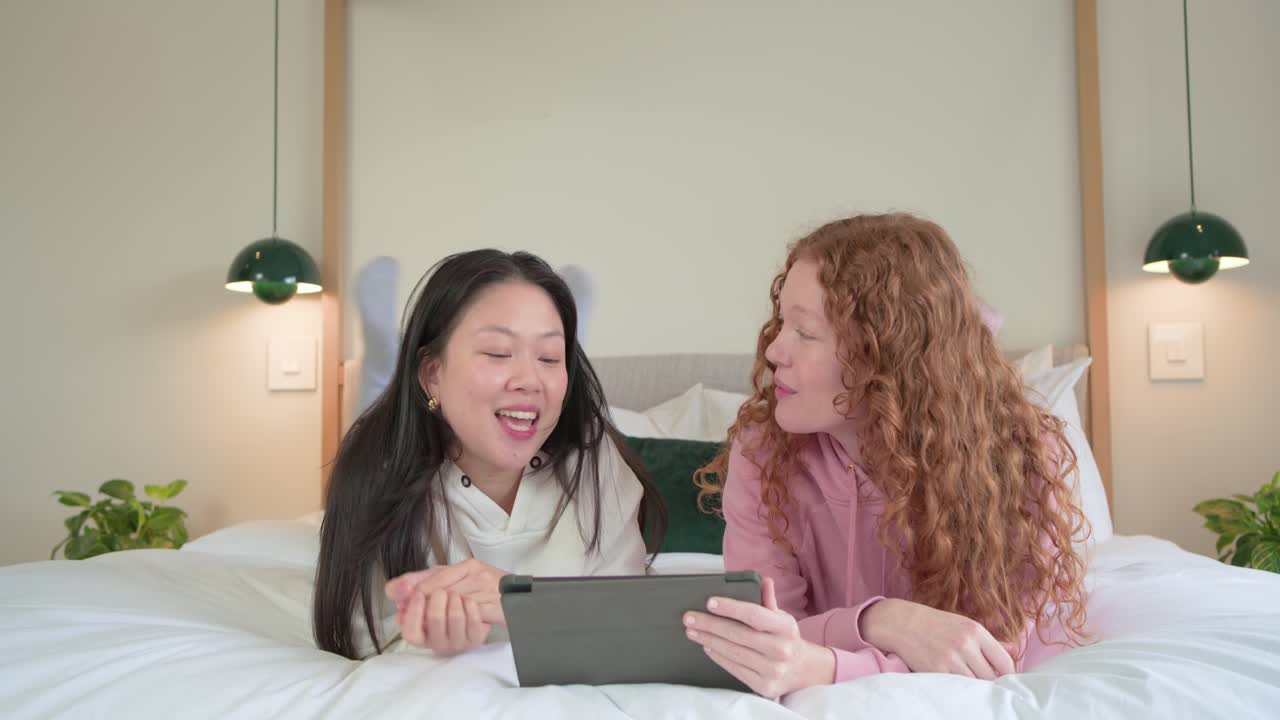Amiche diverse sdraiate sul letto che guardano un clip divertente su un tablet e ridono insieme