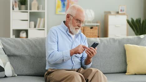 Alter-Grauhaariger-Mann-Mit-Brille,-Der-Zu-Hause-Auf-Der-Couch-Sitzt-Und-Eine-Nachricht-Auf-Dem-Smartphone-Schreibt