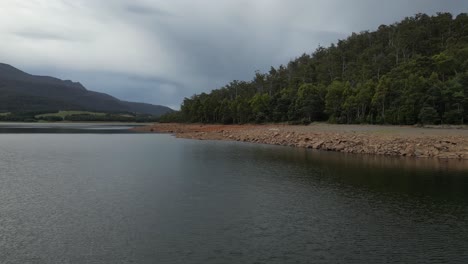 Low-altitude-flight-over-Lake-Huntsman-with-rocky-shore-and-green-forest-hills-at-cloudy-day