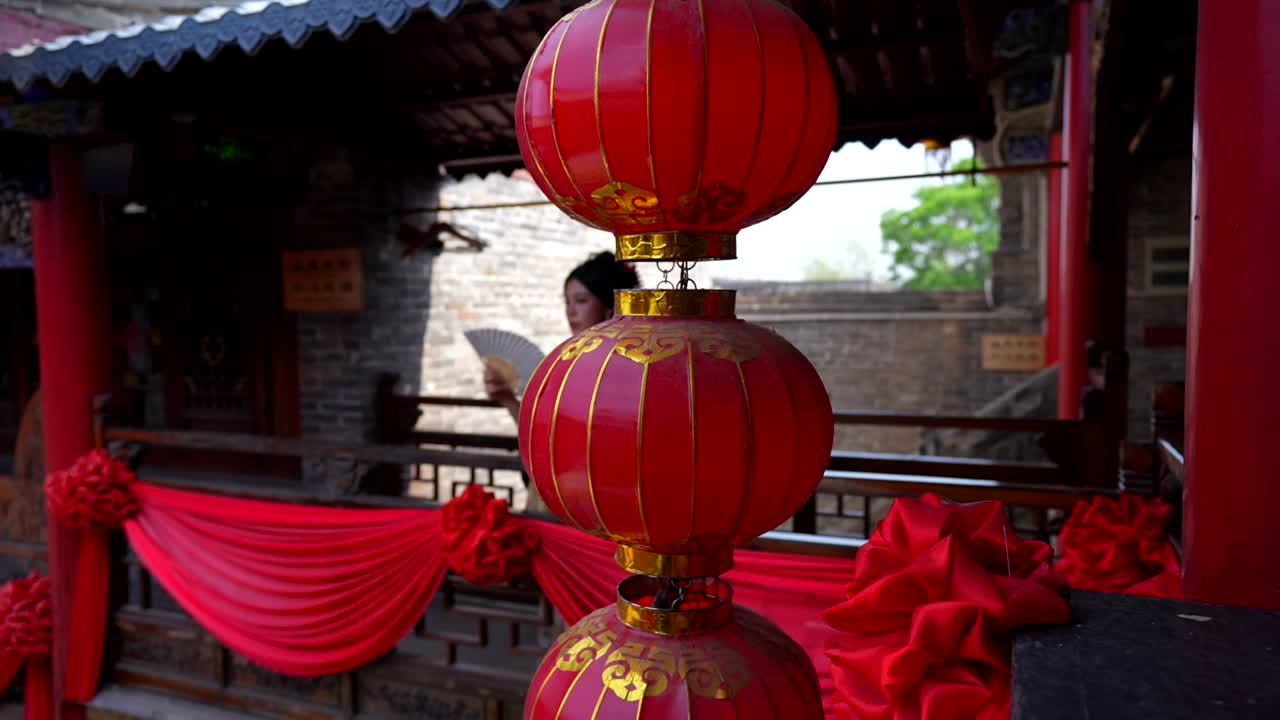 Đèn lồng đỏ ở tiền cảnh, cùng một cô gái mặc trang phục triều Thanh đang quạt mát trên ban công truyền thống Trung Quốc trang trí bằng các vật phẩm màu đỏ.