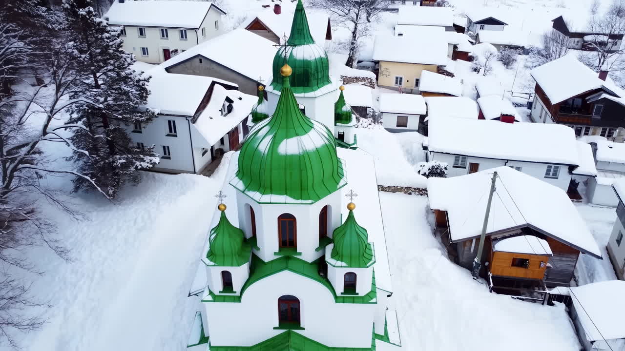 Letecký pohled na kostel se zelenými kupolemi nad zasněženou vesnicí, filmový dron se vznáší nad dřevěnými střechami a úzkými uličkami.