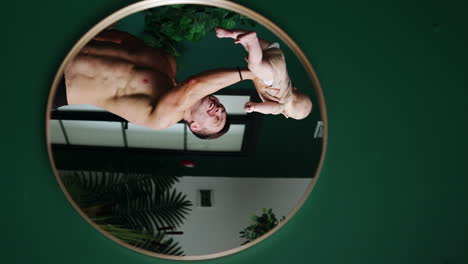sportive dad holding his baby over the head with one hand. reflection in the round mirror on the green wall. vertical screen.