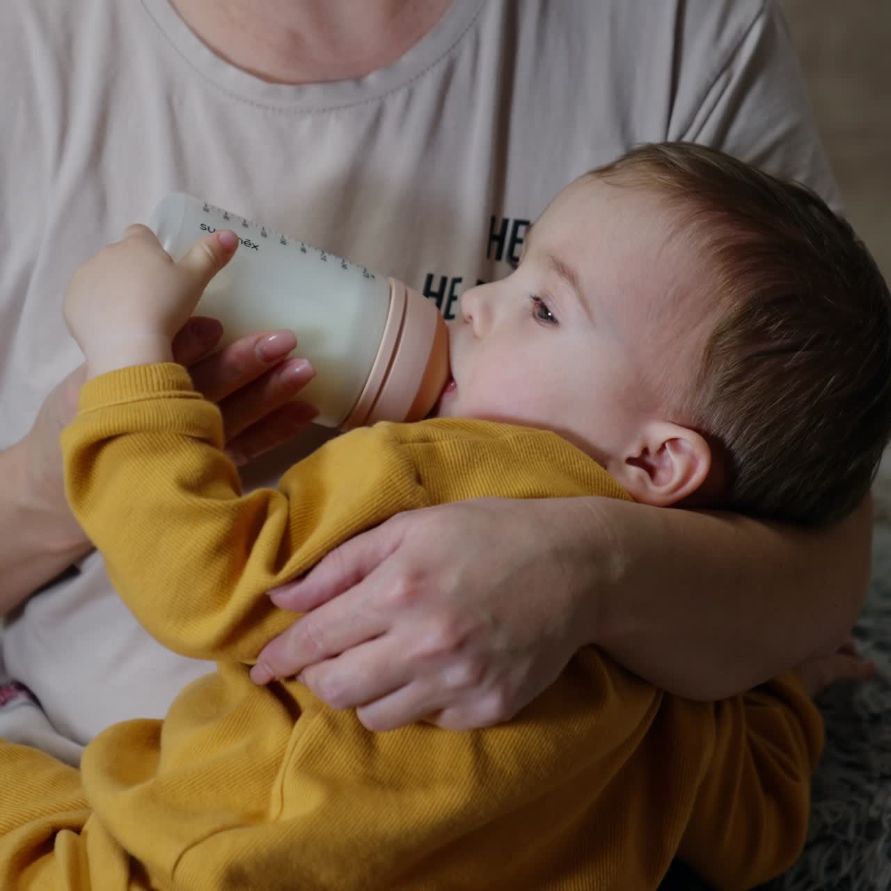 A cute toddler lies in mom's hands eating from bottle. Baby boy supporting the bottle with his hand