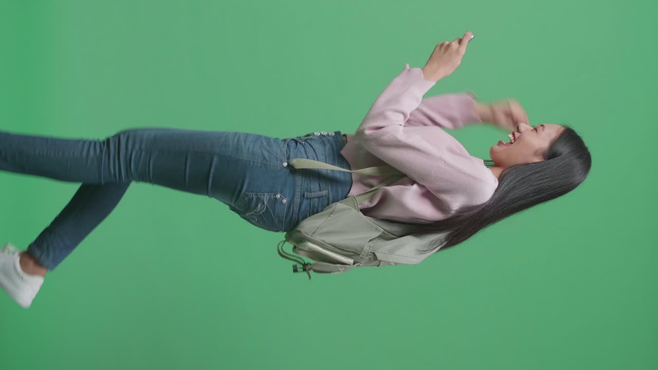 vista lateral de una estudiante asiática feliz jugando a un juego móvil y ganando mientras camina en el estudio de pantalla verde