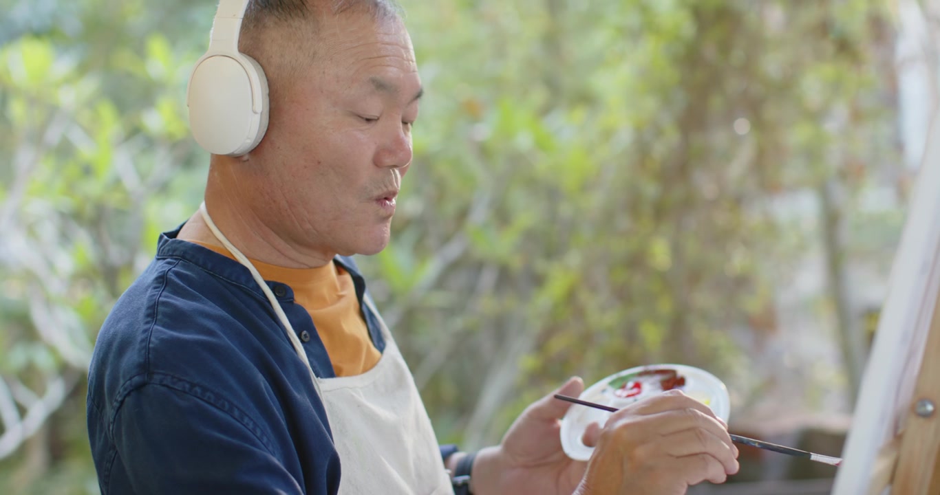 Anziano uomo asiatico che mescola la vernice sulla tavolozza, applicandola con un pennello sulla tela in veranda, creando un'opera d'arte.