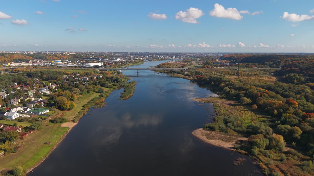 Aerial view of the Nemunas River flowing through Kaunas, Lithuania, showcasing vibrant autumn colors, residential areas, and scenic natural landscapes under a blue sky with scattered clouds