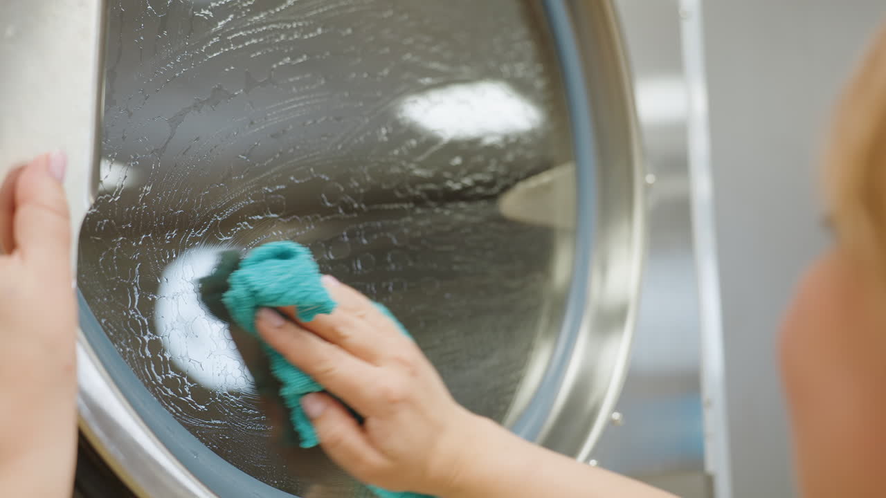 Hand view of elegant woman cleaning transparent surface door of washing machine with soapy towel, leaving visible soap marks, her reflection faintly visible on glass during cleaning process