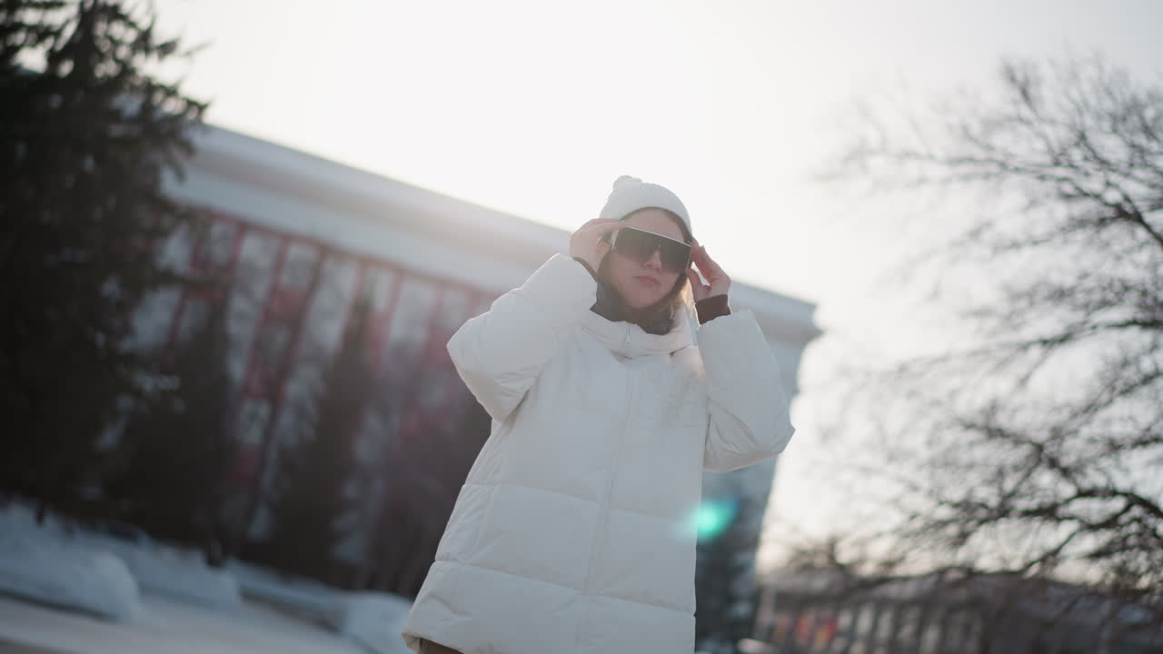 Close up student putting on dark glasses in snowy park, wearing warm winter coat and beanie, rosy cheeks glowing under soft sun, evergreen trees surrounding while adjusting eyewear