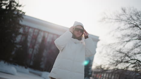 close up student putting on dark glasses in snowy park, wearing warm winter coat and beanie, rosy cheeks glowing under soft sun, evergreen trees surrounding while adjusting eyewear