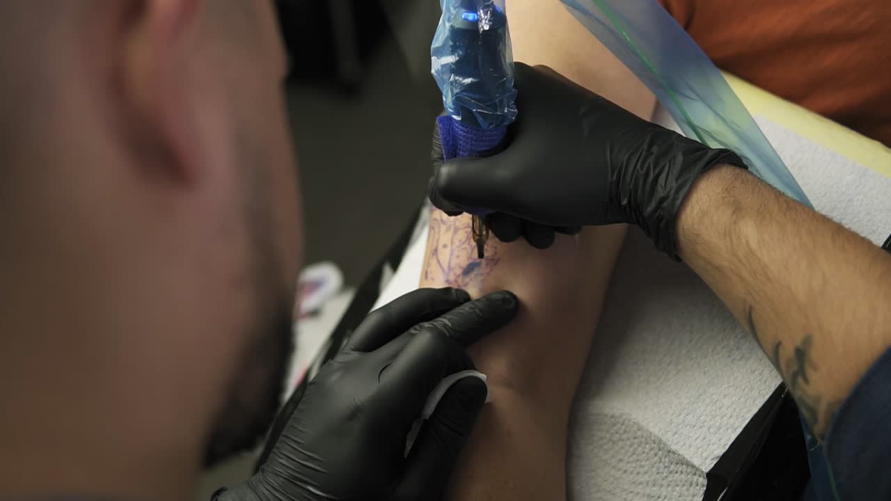A close-up of a tattoo artist makes a tattoo on a woman's arm. A high resolution. Professional tattooist works in studio. High angle footage from the shoulder of master