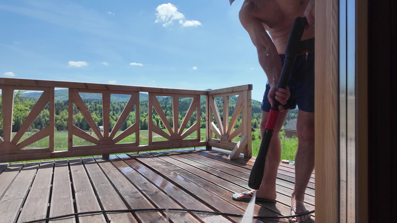 Shirtless man wearing shorts and a cap washes a wooden terrace with a pressure washer at a log cabin under a bright blue summer sky.