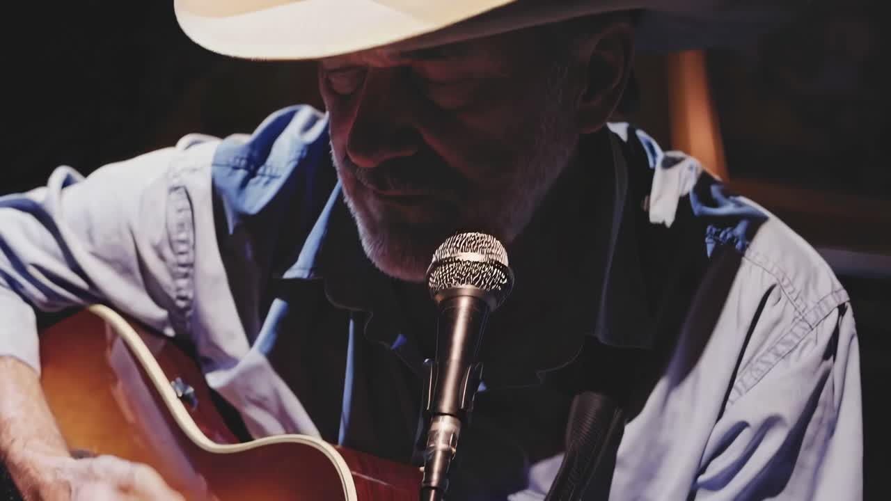 Video ravvicinato di un musicista cowboy che canta in un microfono, con un'illuminazione drammatica in risalto