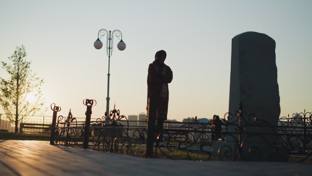 코트와 스카프를 입은 슬픈 여자가 해가 지는 동안 공원에서 돌로 된 기념비 앞에 서 있습니다. 슬픈 자세로, 서있는 태양의 따뜻하고 희미한 빛은 긴 그림자를 던집니다.