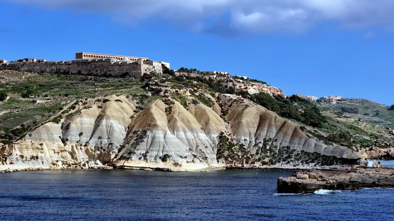 Bredt lavt engel-billede fortæller den historiske historie om Fort Chambray oven på dramatiske stribede lerklinter i Gozo Malta under en lys forårsdag med dybt blåt havvand