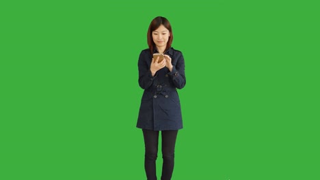 푸른 코트를 입은 젊은 아시아 여성, 스마트폰, 디지털 태블릿을 사용하고 카메라를 보고, 크로마 키를 위해 녹색 화면 앞에 서있는 여성.