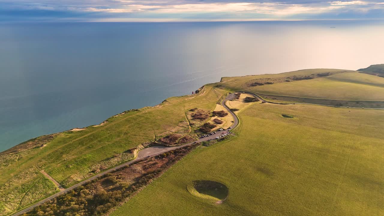 Aerial drone footage over green fields before a cliff-top top-off reveals a bird’s-eye view of Beachy Head’s dramatic white cliffs, iconic lighthouse, rocky beach, and calm turquoise waters.