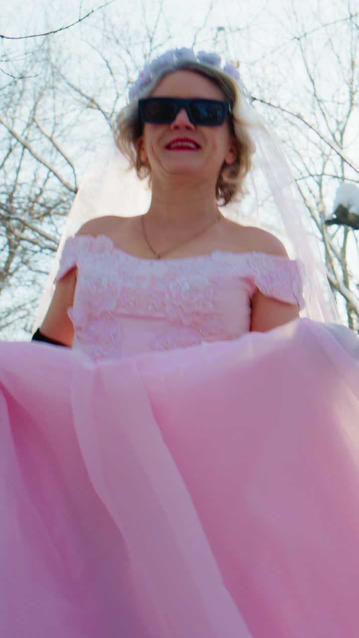 Mujer alegre con vestido de novia rosa y gafas de sol al aire libre