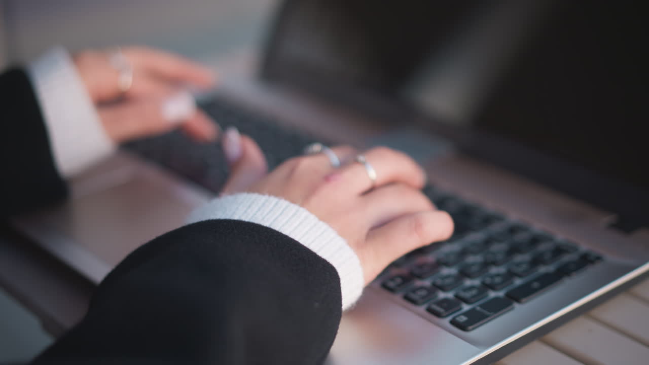 Close-up of hands adorned with polished nails and delicate rings typing on laptop keyboard with slight blur and soft light reflection on screen
